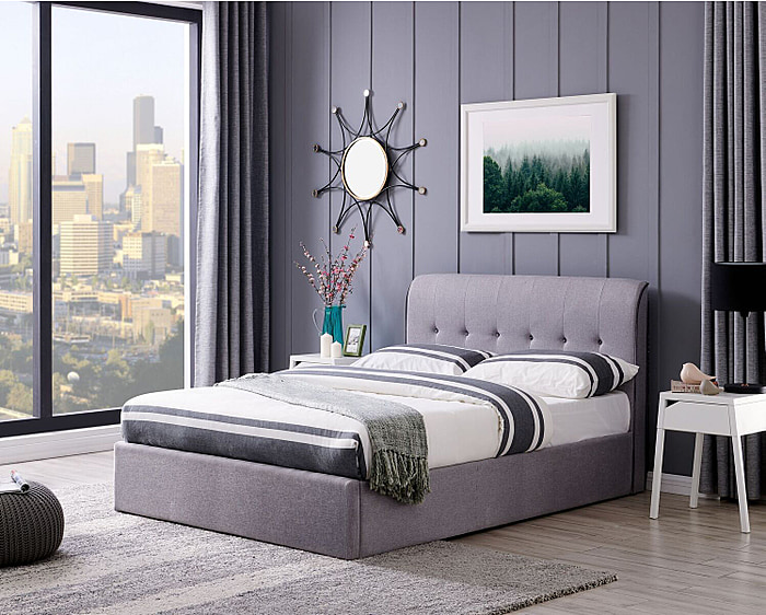 A contemporary, minimalistic grey linen ottoman bed frame. It has a curved headboard with buttons and piping to the edges.